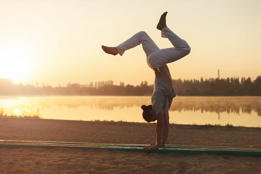 Concept about lifestyle and sport. Capoeira on the beach, near lake one performer, at sunrise. Athletic man in white pants standing on arms upside down while workout in the park in the heart of city. - A Natural Way to Detox Your Body