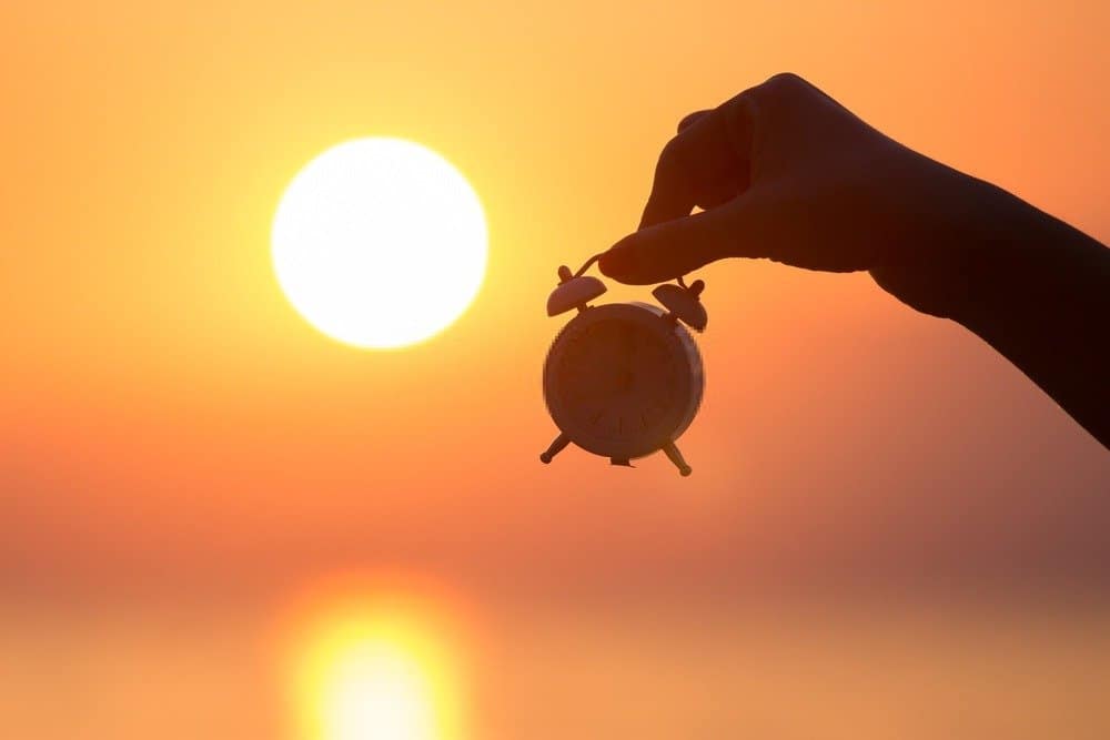 Silhouette of female hand holding small alarm clock and sunrise over sea in the background. Getting up early concept - Start Early