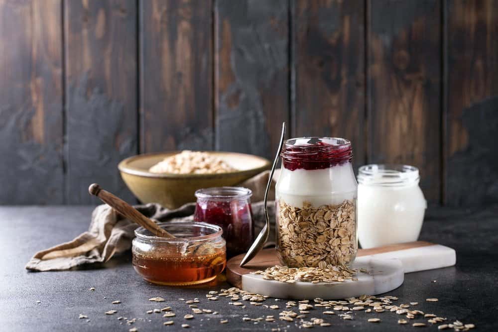 Dry oats served with fresh greek yogurt, strawberry jam and thyme honey, decorated with spiral ornament and honey dipper over a grey background - The Metabolic Reset Diet Plan