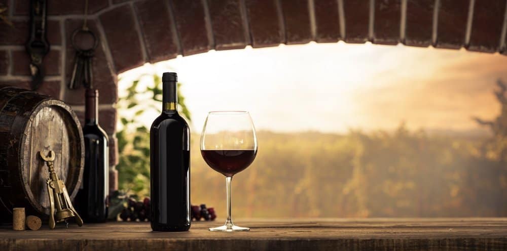 Red wine tasting in the wine cellar: wineglass and bottles next to the window and panoramic view of vineyards at sunset