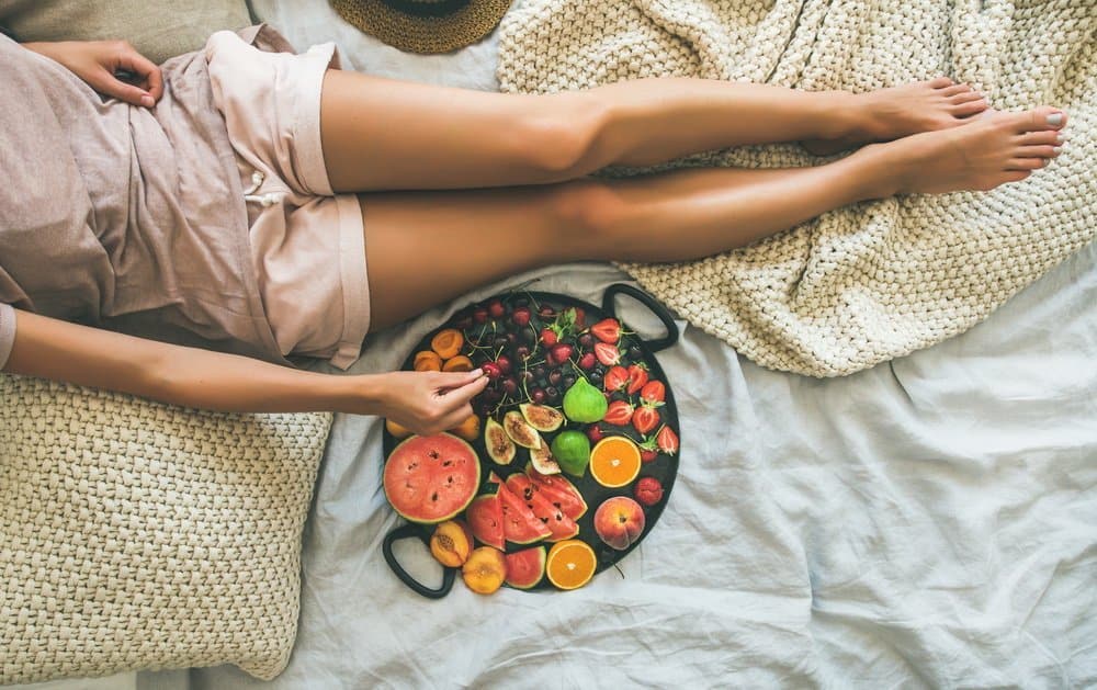 Summer healthy raw vegan clean eating breakfast in bed concept. Young girl wearing pastel colored home clothes taking fruit from tray full of fresh seasonal fruit. Top view, copy space - A Natural Way to Detox Your Body