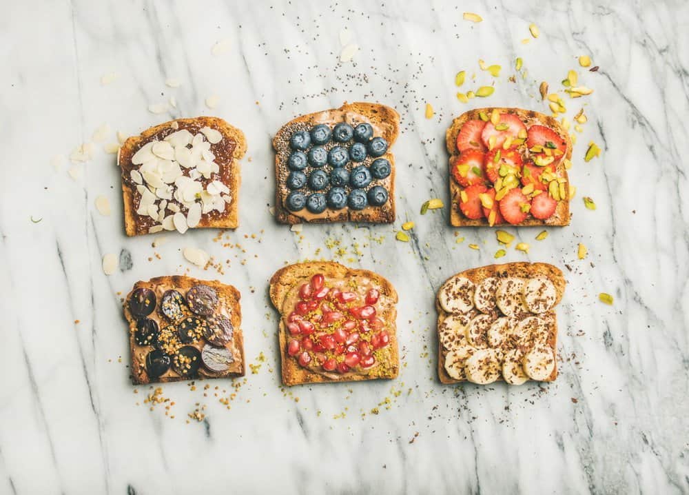Healthy breakfast or snack. Flat-lay of vegan whole grain toasts with fruit, seeds, nuts and peanut butter over marble background, top view. Clean eating, vegetarian, dieting food concept