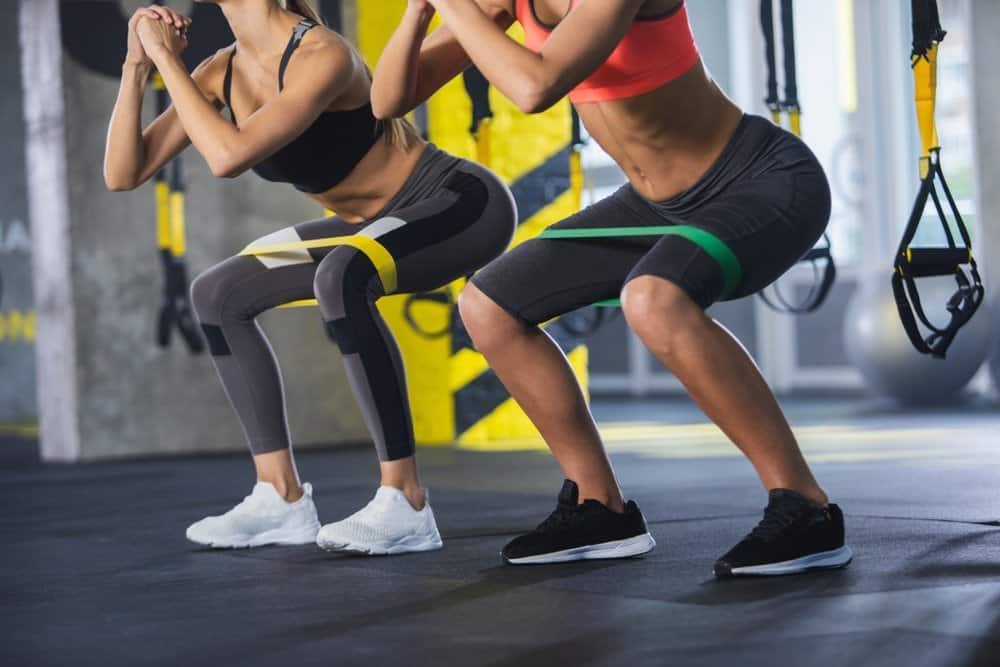 Close up of athletic women in squat together in gym. Couple of fit girls are exercising - Commit to workout with a close friend