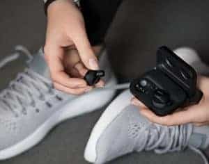 Person holding black noise cancelling wireless earbuds next to open charging case, with gray sneakers visible in the background, showcasing best earbuds for music in 2025.