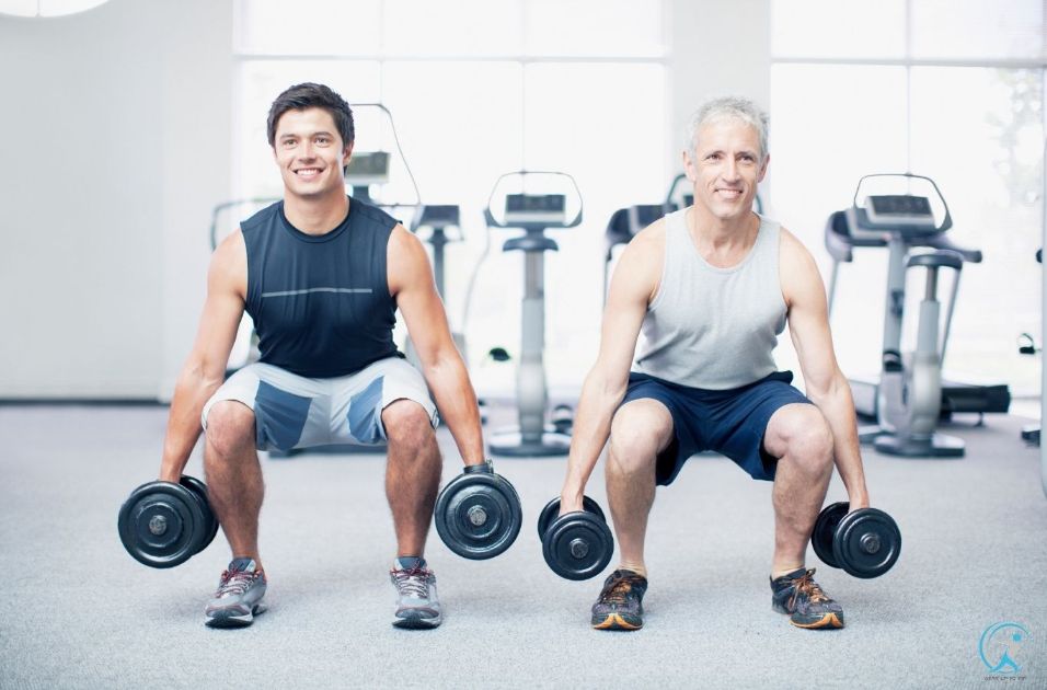 A young and an older athlete are performing squats with dumbbells
