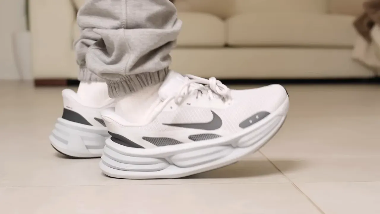 Person wearing white Nike running shoes with gray accents, standing on a tiled floor.