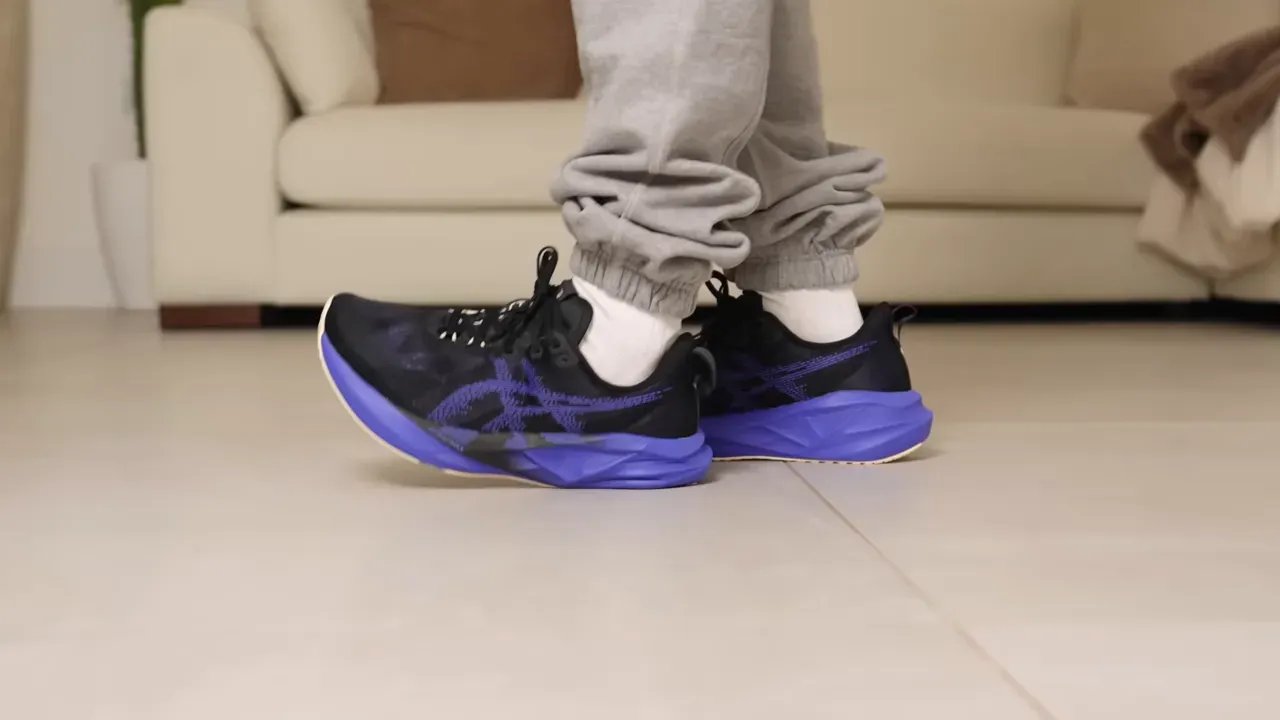 Person wearing black and purple ASICS running shoes, standing on a tiled floor.