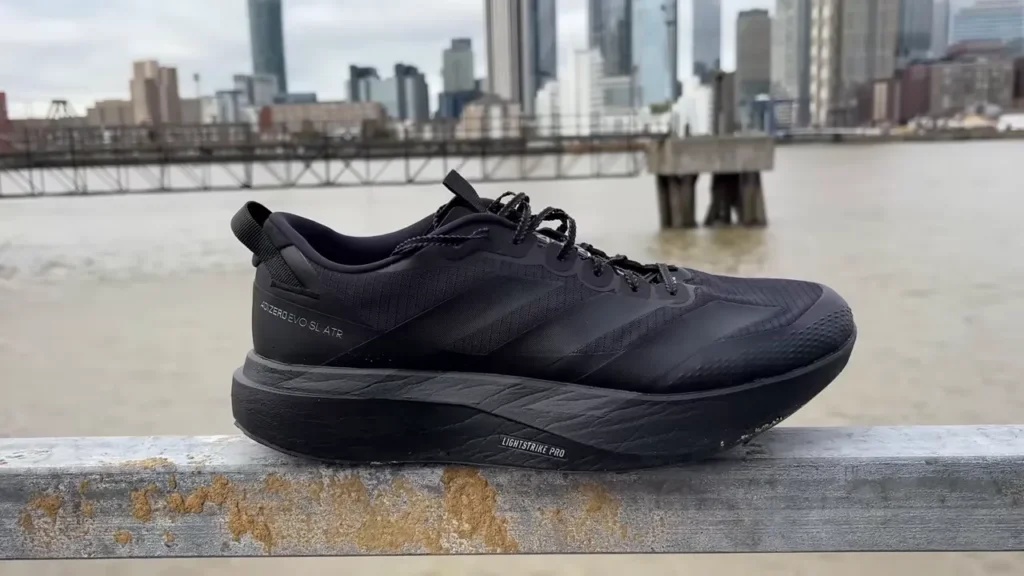 Adidas EVO SL ATR shoe in black, resting on a railing with a city skyline in the background.